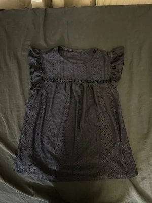 Svart topp med volangärmar - Snygg svart topp med korta volangärmar och broderat hålmönster. Rund halsringning och dekorativ spetsdetalj under bysten. Luftig och lätt i materialet, perfekt för en trendig look.