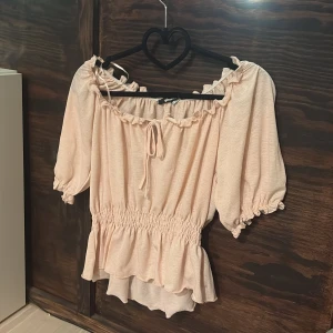 Ljusrosa blus från Gina Tricot - Supersöt ljusrosa blus från Gina Tricot med volangkant, smock i midjan och puffiga korta ärmar. Blusen har en rundad halsringning som är stretchig med knyt och rynkade detaljer som ger en romantisk vibe. Perfekt att styla med jeans eller kjol! Använt en gång då den inte kommer till användning längre!