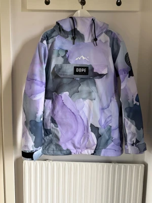 DOPE skidjacka  - Säljer en unik skidjacka/snowboardjacka från Dope med abstrakt mönster i lila, grått och vitt. Jackan har huva och stor magficka med Dope-logga och kardborre vid ärmsluten. Perfekt för dig som vill sticka ut med en färgstark och modern look i skidbacken. Nypris: 2099kr.