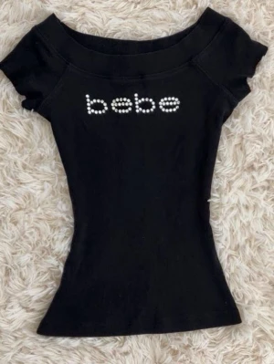 Svart topp från Bebe med pärldetaljer - Svart figurnära topp från Bebe med korta ärmar och pärlprydd logga framtill. Snygg, enkel och perfekt att styla med jeans eller kjol för en trendig look.