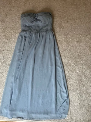 Ljusblå axelbandslös långklänning Vila - Säljer en ljusblå axelbandslös långklänning från Vila. Klänningen har ett vackert flytande, lätt tyg som ger en elegant siluett. Perfekt för sommarens tillfällen och studentfirande.