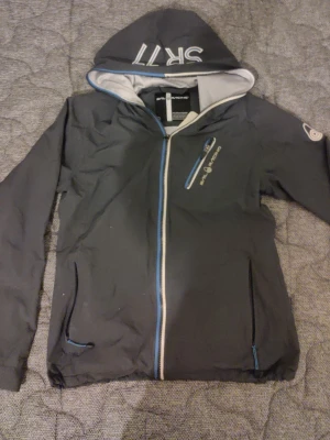 Grå/blå vindjacka från Sail Racing - Grå/blåvindjacka från Sail Racing med huva och vita detaljer. Jackan har blå och vit dragkedja, två sidofickor och en bröstficka med dragkedja. Innerfodret är i mesh och huvan har trycket 'SR11'. Perfekt för blåsiga dagar.
