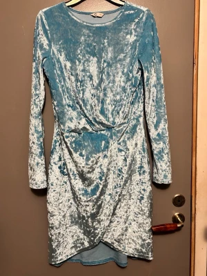 Blå sammetsklänning från Cubus - Snygg blå sammetsklänning med omlott-detalj framtill och långa ärmar. Klänningen har en kort längd och en glansig, mjuk yta som ger en lyxig känsla. Perfekt för dig som vill sticka ut med en unik look.