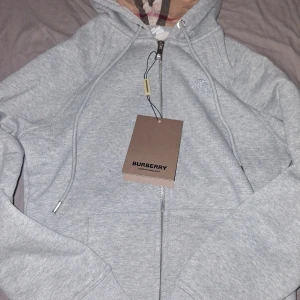 Grå hoodie med dragkedja, till man men funkar i unisex, pris kan diskuteras - Snygg grå hoodie med dragkedja framtill och klassiskt rutigt foder i huvan. Broderad logga på bröstet och fickor på sidorna. Tillverkad i mjuk bomullsblandning, perfekt för en avslappnad och trendig look.