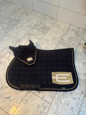 Svart schabrak och huva Equiline - Säljer ett svart schabrak och matchande huva från Equiline. Båda har guldfärgade detaljer och en stor vit logga. Materialet är quiltat tyg med kantband i guld. Perfekt för dig som vill ha en snygg och stilren look till hästen. BRA SKICK