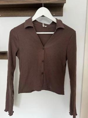 Brun ribbad cardigan med krage - Snygg brun ribbad cardigan med krage och knäppning framtill. Långärmad och croppad modell som passar perfekt till jeans eller kjol. Mjuk och stretchig i materialet för en bekväm känsla.