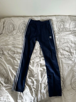 Mörkblå Adidas mjukisbyxor - Säljer ett par mörkblå mjukisbyxor från Adidas med klassiska vita ränder längs sidorna och logga på låret. Byxorna har en slim fit passform och resår vid benslut. Perfekta för chill eller träning.