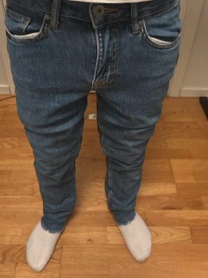 Blå raka jeans med vita detaljer - Säljer ett par klassiska blå jeans med raka ben och vita detaljer vid fickorna. Jeansen har fem fickor, bälteshällor och knappgylf. Perfekta för en avslappnad och stilren look. Materialet är denim i bomull.