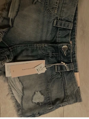 Blå denimshorts med slitningar - Säljer ett par blå denimshorts från Gina Tricot med råa kanter och slitna detaljer. Shortsen har hög midja, klassiska fem fickor och knappgylf. Perfekta för sommaren och ger en avslappnad look.