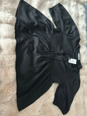 Svart blus med knytband från lager 157 - Stilren svart blus från lager 157 med lätt glansigt tyg och struktur. Blusen har korta ärmar och knytband framtill vid halsen. Passar perfekt till jeans eller kjol för en trendig look.  Storlek:M