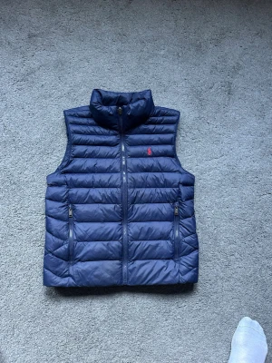 Mörkblå dunväst från Ralph Lauren - Snygg mörkblå dunväst från Ralph Lauren med horisontella sömmar och hög krage. Västen har två dragkedjefickor framtill och en liten röd logga på bröstet. Perfekt för lager-på-lager och chill stil.