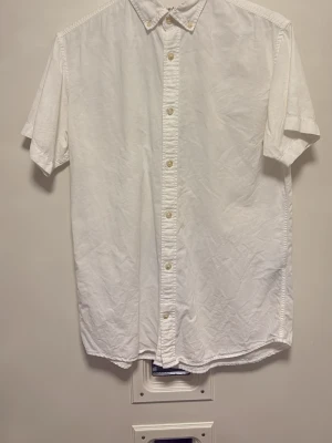 Vit kortärmad skjorta med knappar - Klassisk vit skjorta med korta ärmar och knappar hela vägen framtill. Skjortan har en enkel och stilren design med krage och är perfekt för en avslappnad men snygg look.