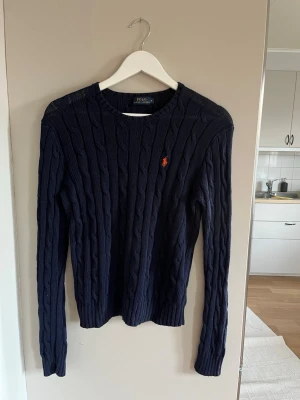 Mörkblå kabelstickad tröja Polo Ralph Lauren - Säljer en mörkblå kabelstickad tröja från Polo Ralph Lauren med den klassiska orange broderade loggan på bröstet. Tröjan har rund halsringning, långa ärmar och ribbade muddar. Perfekt för dig som gillar stilrena och tidlösa plagg.