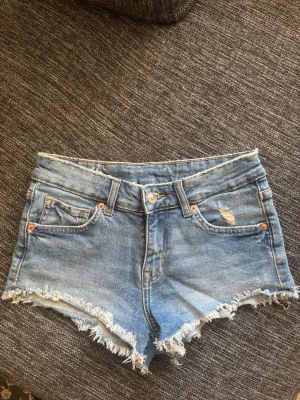 Blå jeansshorts med fransig kant - Snygga blå jeansshorts med fransig kant och slitna detaljer. Klassisk femficksmodell med knapp och dragkedja framtill. Perfekta för varma dagar och enkel att matcha med allt. Storlek 34💞