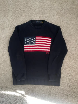 Mörkblå stickad tröja Polo Ralph Lauren - Mörkblå stickad tröja från Polo Ralph Lauren med amerikansk flagga i rött, vitt och blått på bröstet. Rund halsringning och ribbstickade muddar vid ärmslut och nederkant. Klassisk och stilren design med RL-broderi.