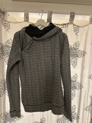 Gråmelerad hoodie med stor huva - Säljer en gråmelerad hoodie med stor, mysig huva och långa ärmar. Tröjan har en cool asymmetrisk krage och är gjord i ett mjukt, stickat material. Perfekt för dig som gillar en avslappnad och stilren look.