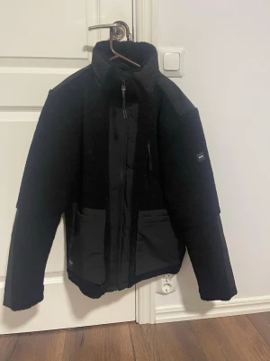 Svart teddyjacka  - Säljer en svart teddyjacka från Revolution med detaljer i nylon på bröst, axlar och fickor. Jackan har hög krage, dragkedja framtill, två stora framfickor och en bröstficka med dragkedja. Perfekt för dig som vill ha en stilren och mysig jacka.