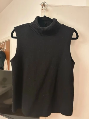 Svart ärmlös stickad polotröja Zara - Svart stickad ärmlös polotröja från Zara. Tröjan har en hög, ribbad polokrage och är gjord i ett mjukt material som känns skönt mot huden. Perfekt att styla lager på lager eller bära som den är för en clean look.