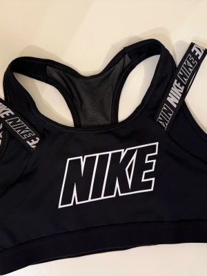 Svart sport-bh från Nike - -Sport-bh från Nike-breda axelband med upprepande Nike-logotyp. -Materialet är stretchigt och har meshdetaljer i ryggen för extra ventilation.  -Använd kanske 2 gånger och tvättad. Som ny -Ordinarie pris: 599 kr -Perfekt för träning och aktiviteter.
