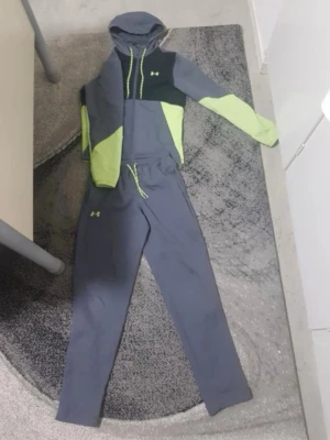 Under Armour grå och limegrön loungewear - Mysig loungewear-set från Under Armour i grått med limegröna detaljer. Setet består av en hoodie med dragsko och matchande byxor med resår och snörning i midjan. Perfekt för chill dagar hemma eller när du vill vara bekväm.