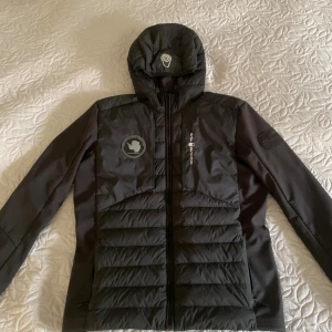 Sail racing hybrid  - Svart dunjacka/hybrid från Sail Racing med huva och snygga logodetaljer på bröstet och ärmen. Jackan har quiltad framsida och baksida, dragkedja hela vägen och är tillverkad i slitstarkt syntetmaterial med fyllning av dun (800 fill power). Perfekt för kalla dagar. Köpte för ny pris 3500kr