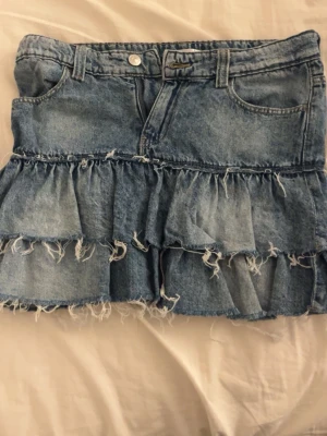 Blå jeanskjol med volanger - Trendig blå jeanskjol med två volanglager och fransiga kanter. Kjolen har klassiska jeansdetaljer som fickor fram och bak samt bälteshällor. Perfekt för dig som gillar en edgy och avslappnad stil.