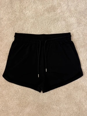 Svarta shorts med snörning - Svarta shorts med elastisk midja och snörning framtill. De har en enkel och sportig design med mjukt material som känns skönt mot huden. Perfekta för varma dagar eller när du vill chilla hemma.