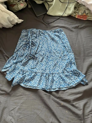 Blå blommig kortkjol med resår - Säljer en blå kortkjol med vitt blommigt mönster och resår i midjan. Kjolen har en luftig och lätt form, perfekt för varma dagar. Materialet känns mjukt och bekvämt mot huden. Hålet ser större ut än mad det är i en söm men det är lätt att sy ihop.