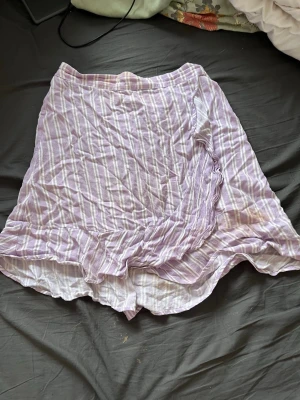 Ljuslila randig omlottkjol - Söt ljuslila kortkjol med vita ränder och omlottdetalj. Kjolen har volang längs nederkanten och är gjord i ett lätt och luftigt bomullstyg, perfekt för varmare dagar. Resår i midjan för skön passform. Det finns lite sminkfläckar på den men det går bort i tvätten eller med sminkborttagning.