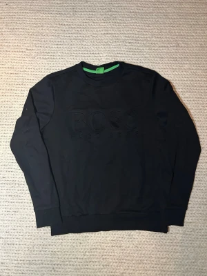Svart sweatshirt från Hugo Boss - Säljer en svart sweatshirt från Hugo Boss med diskret broderad logga framtill. Tröjan har rund halsringning, långa ärmar och ribbade muddar. Tillverkad i mjuk bomull för skön känsla och stilren look.