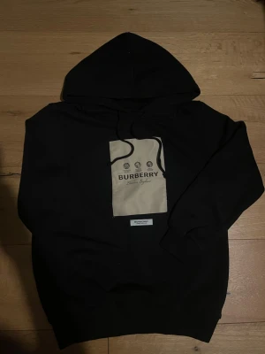Svart hoodie från Burberry - Snygg svart hoodie från Burberry med stor beige patch och logga på bröstet. Klassisk huva med dragsko och ribbade muddar vid ärmslut och nederkant. Tillverkad i mjukt material som känns skönt mot huden. Perfekt för en trendig och avslappnad stil. Jag möts bara upp om man vill köpa.