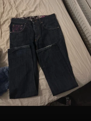 Mörkblå jeans med lila detaljer - Säljer ett par mörkblå jeans med raka ben och unika lila detaljer på fickorna och bakfickorna. Jeansen har en cool patch med lila tryck och kontrastsömmar. Perfekta för dig som vill sticka ut lite extra i vardagen.