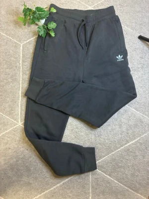 Svarta mjukisbyxor från Adidas - Säljer ett par svarta mjukisbyxor från Adidas med dragsko i midjan och ribbade muddar vid bensluten. Byxorna har två sidofickor och den klassiska Adidas-loggan broderad på vänster sida. Perfekta för chill dagar eller träning.