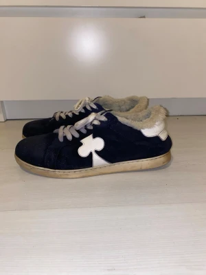 The gallerist moritz - Mörkblåa sneakers med vit sula och vit fuskpäls på insidan. Skorna har en stor vit klöverdetalj på sidan och vita skosnören. Ovandelen är i mocka . Perfekta för dig som vill ha något unikt och mysigt på fötterna.
