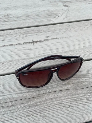 Svarta solglasögon från Vestimo - Snygga svarta solglasögon från Vestimo med rektangulära bågar och mörkröda glas. Klassisk design med diskreta silvriga detaljer vid sidorna. Perfekta för att ge din stil en cool touch under soliga dagar.