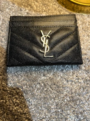Svart YSL korthållare i läder - Snygg svart korthållare från Saint Laurent i präglat läder med silverfärgad YSL-logga framtill. Stilren design med flera kortfack och diskret mönster. Perfekt accessoar för dig som gillar lyxiga detaljer.