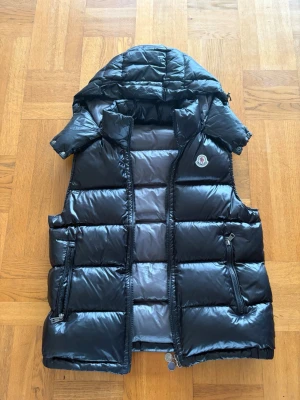 Svart dunväst från Moncler med huva - Snygg svart dunväst från Moncler med avtagbar huva och två dragkedjefickor. Västen har en glansig finish och klassisk Moncler-logga på bröstet. Fodrad med dun för extra värme och komfort. Storlek 2 vilket motsvarar M. Pris går att diskutera!🤩