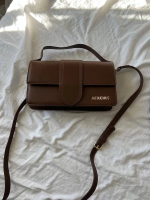 Brun axelväska från Jacquemus - Snygg brun axelväska från Jacquemus i slätt läder med justerbar axelrem och guldfärgad logga framtill. Väskan har en stilren, rektangulär form och stängs med ett lock. Perfekt för dig som gillar minimalistisk design och trendiga accessoarer. Osäker på äkthet därav billigt pris