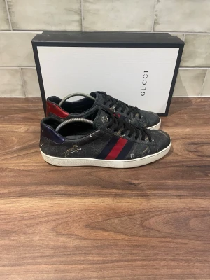Gucci svarta sneakers med röd/blå rand - Gucci sneakers i svart med klassiskt monogrammönster och guldfärgade detaljer. Sidan har en röd och blå rand, och hälen är dekorerad med metallicrött och blått. Vit sula och svarta skosnören. Snygg och exklusiv design som sticker ut.
