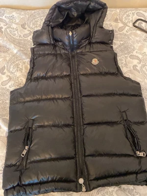 Svart dunväst från Moncler med huva - Snygg svart dunväst från Moncler med avtagbar huva och två dragkedjefickor framtill. Västen har Moncler-logga på bröstet och är tillverkad i glansigt material. Perfekt för lager-på-lager och streetstyle.