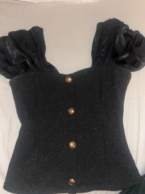 Svart glittrig korsettopp med puffärm - Supersnygg svart korsettopp med glittrigt tyg och stora puffärmar. Toppen har dekorativa guldknappar framtill och en figurnära passform som ger en riktigt trendig look. Perfekt för dig som vill sticka ut lite extra. Använd 1 gång 