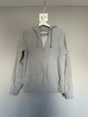 Grå zip-hoodie   - En stilren grå hoodie med dragkedja framtill och huva med dragsko. Tröjan har långa ärmar, ribbade muddar och två fickor på sidorna. Perfekt för en avslappnad look och enkel att matcha med andra plagg.