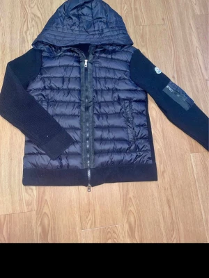 Svart pufferjacka med huva Moncler - Snygg svart pufferjacka från Moncler med stickade ärmar och huva. Jackan har två sidofickor med dragkedja och en Moncler-logga på ena ärmen. Kroppen är quiltad och glansig medan ärmarna är i mjukt stickat material. Perfekt för kalla vinterdagar. Storlek L men sitter som s 