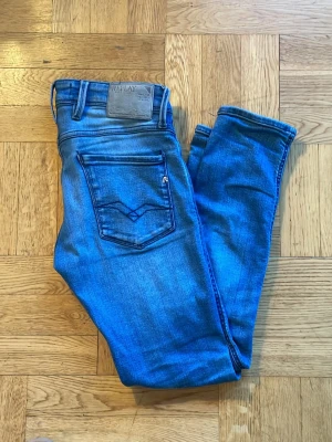  Replay Jeans - Säljer ett par blå slim jeans från Replay med klassisk femficksdesign. Jeansen har en läderpatch med logga bak i midjan och är tillverkade i stretchigt denim för skön passform. Perfekta för dig som gillar en smal siluett. Ett litet hål förekommer på framsidan av jeansen vilket förklarar det billiga priset. Skriv om du har frågor!