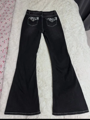 Svarta bootcut jeans med brodyr - Säljer ett par svarta bootcut jeans med vita kontrastsömmar och snygg broderad detalj på bakfickorna. Jeansen har klassisk femficksmodell och utsvängda ben. Perfekta för dig som gillar retrovibbar och vill sticka ut lite extra och högmidjade jeans. 