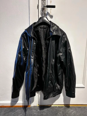 Svart klassisk skinnjacka - Snygg svart skinnjacka med bred krage och dragkedja framtill. Jackan har en avslappnad passform, elastisk nederkant och muddar vid ärmsluten. Perfekt för dig som vill ha en tidlös och cool look.