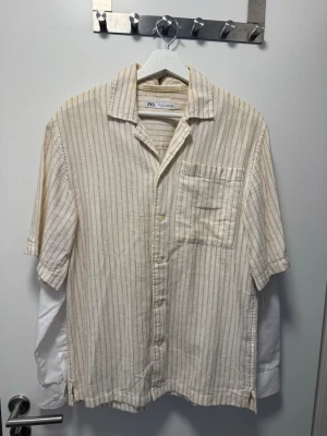 Beige randig skjorta från Zara - Passer även M. Säljer en beige kortärmad skjorta från Zara med tunna bruna ränder och öppen krage. Skjortan har en bröstficka och knäppning framtill. Tillverkad i ett mjukt linnematerial som passar perfekt till varmare dagar.