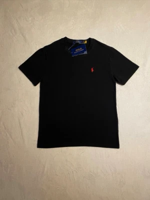 Svart t-shirt från Polo Ralph Lauren - Svart t-shirt från Polo Ralph Lauren med klassisk passform och rund hals. T-shirten har en röd broderad logga på bröstet och är tillverkad i mjuk bomull. Perfekt för en stilren och enkel look.