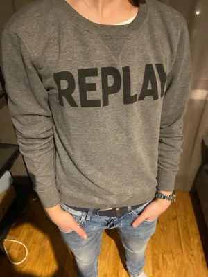 Grå sweatshirt från Replay - Säljer en grå sweatshirt från Replay med stor svart logga över bröstet. Tröjan har rund halsringning, långa ärmar och ribbade muddar vid ärmslut och nederkant. Perfekt för en avslappnad och stilren look.
