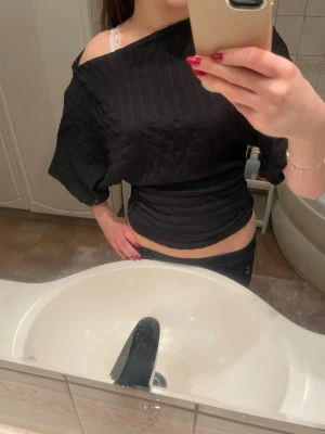 Svart offshoulder topp med struktur - Trendig svart offshoulder topp med strukturerat tyg och croppad passform. Toppen har vida ärmar och sitter tight över midjan för en snygg siluett. Perfekt för dig som vill ha en unik och stilren look.